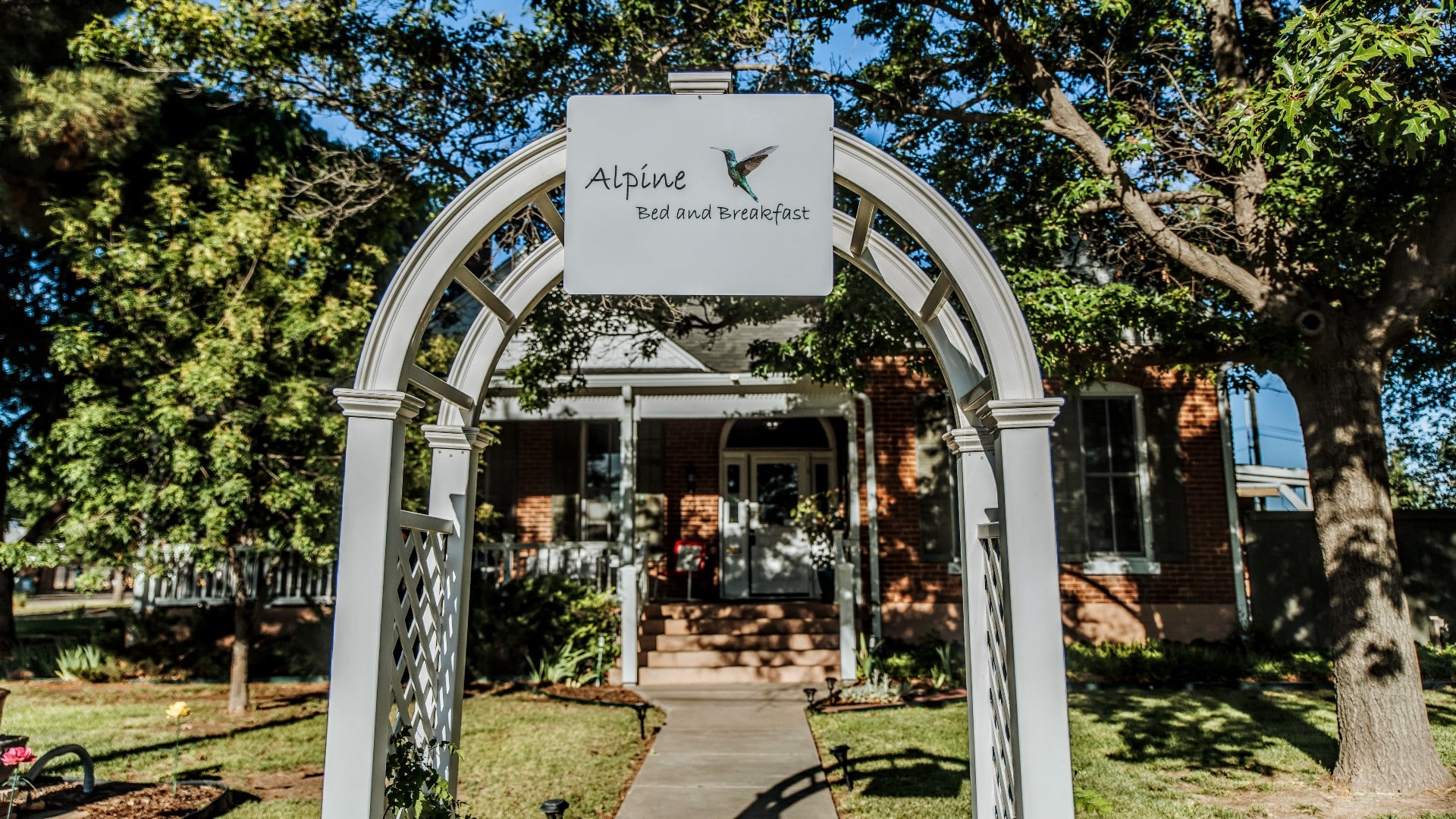 An archway with a business sign at the end of a sidewalk in front of a brick home surrounded by trees.