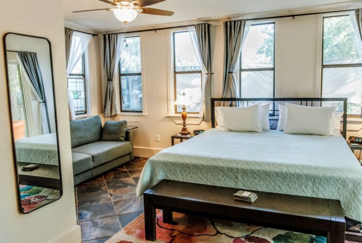 A large bedroom with five large windows, king bed, side tables with lamps and loveseat