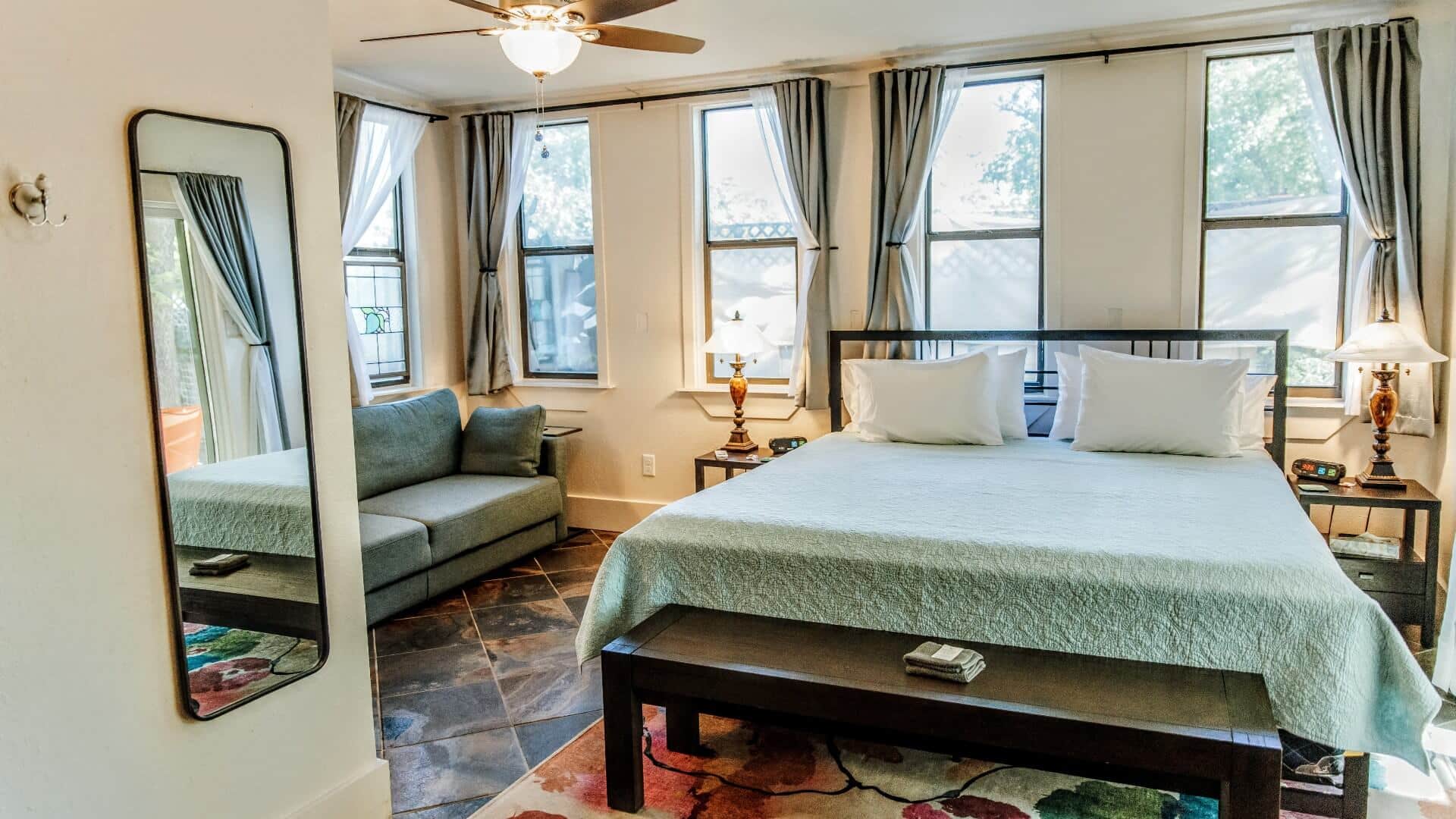 A large bedroom with five large windows, king bed, side tables with lamps and loveseat