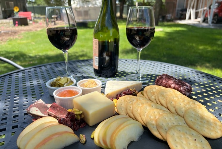 A charcuterie platter with fruit, crackers, meats and cheeses on an outdoor table with two wine glasses and a bottle of wine