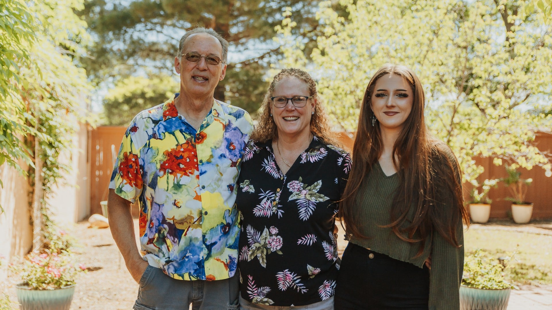 A man, woman and young girl standing close for a photo outside surrounded by trees and sunshine