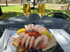 12 shrimp and 2 lemon wedges on the side of a bowl filled with red sauce, on a plate with crackers. Two glasses of bubbly rose wine are behind the plate.
