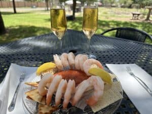 12 shrimp and 2 lemon wedges on the side of a bowl filled with red sauce, on a plate with crackers. Two glasses of bubbly rose wine are behind the plate.