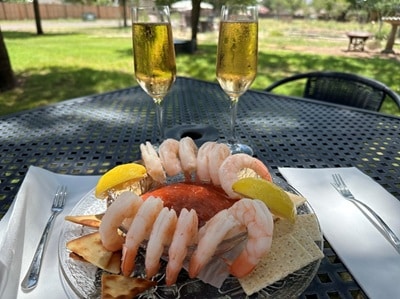 12 shrimp and 2 lemon wedges on the side of a bowl filled with red sauce, on a plate with crackers. Two glasses of bubbly rose wine are behind the plate.