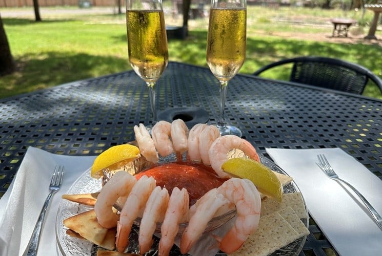 12 shrimp and 2 lemon wedges on the side of a bowl filled with red sauce, on a plate with crackers. Two glasses of bubbly rose wine are behind the plate.